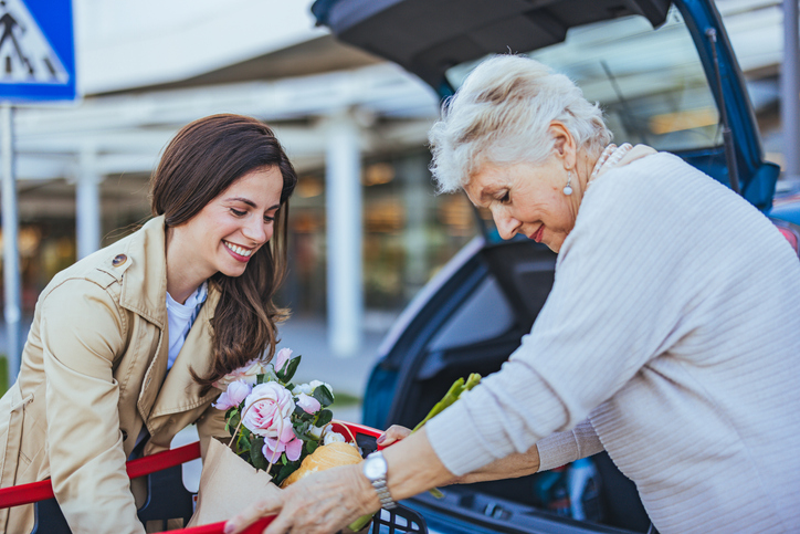 Eine Alltagsbetreuerin begleitet eine hilfebedürftige Dame beim Einkauf und hilft ihr, die Sachen in den Kofferraum des PKWs zu räumen. Die Alltagsbetreuerin zeigt ein freundliches Lächeln, während sie die Dame unterstützt, um ihr mehr Unabhängigkeit und Selbstständigkeit zu geben.